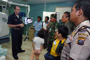 Eine Gruppe von Menschen steht in einem Krankenhauszimmer, ein Mann hält ein Mikrofon in der Mitte, ein Bett mit einer Decke darauf links und verschiedene medizinische Geräte im Hintergrund.
