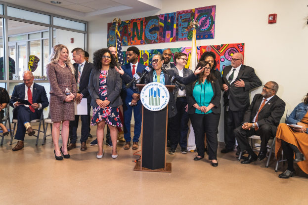 Gruppe von Menschen vor einem Rednerpult mit Mikrofon und einer Tafel mit der Aufschrift "Sancho City Councilman Joins City Council", mit sitzenden und stehenden Gästen, Flaggen, Feuerlöscher und Deckenleuchten im Hintergrund.