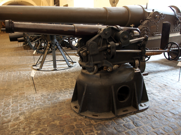 Eine große Gatling-Kanone mit langem Lauf und Seitengriff ist in einem Museum ausgestellt, umgeben von Bodenständen, mit einer fensterlosen Wand im Hintergrund, die an eine Burg erinnert.
