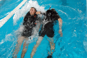 Zwei Personen in schwarzen Neoprenanzügen und Tauchausrüstung befinden sich in einem Schwimmbad, eine mit einem Sauerstoffzylinder auf dem Kopf, beide führen einen Rückenschwimmzug mit ausgestreckten Armen und Beinen aus.