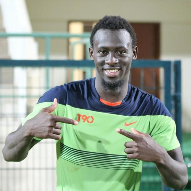 Ein Mann in einem grünen und blauen Hemd lächelt und zeigt auf die Kamera, mit einem unscharfen Geländer, einer Wand und einer Tür im Hintergrund.