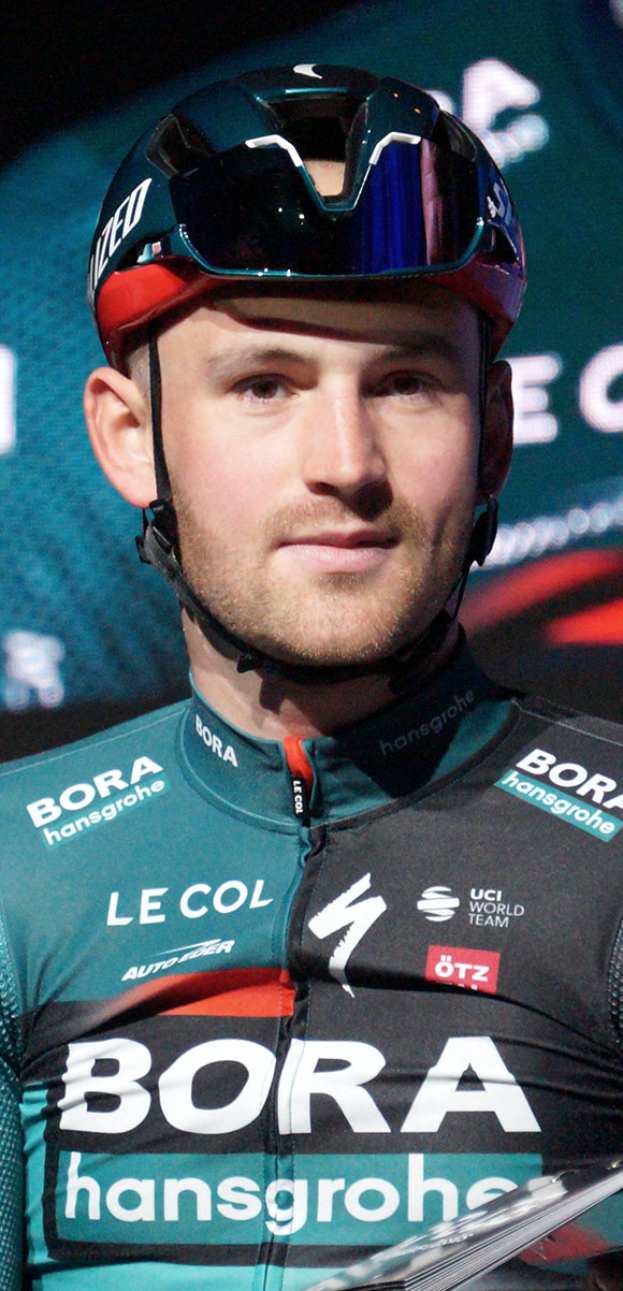 Ein Mann in einem blauen Helm und Sonnenbrille steht neben einem Fahrrad, trägt ein Trikot mit dem Bora-Hansgrohe World Tour-Logo und sieht entschlossen aus.