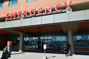 Eine Gruppe von Menschen, die vor einem Notrufgebäude mit Glaswänden, Säulen, einer Namensplakette, Schildern auf Pfählen und einem Mülleimer gehen.