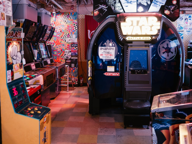 Ein hell erleuchteter Arcade-Raum mit mehreren Maschinen, darunter ein Star Wars Battle Pod, farbenfrohe Poster an den Wänden und verstreute Gegenstände, die eine lebendige Atmosphäre schaffen.