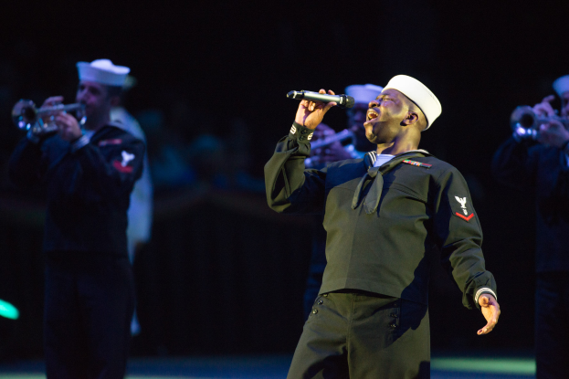 Ein Mann in einer Matrosenuniform singt in ein Mikrofon, umgeben von einer Gruppe von Menschen mit Mützen und Musikinstrumenten, vor einem dunklen Hintergrund, der eine dramatische Atmosphäre schafft.