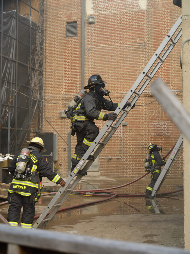 Feuerwehrleute in Helmen und Lufttanks klettern an einer Leiter vor einem Backsteingebäude mit Rohren auf dem Boden und einer Metallstange am Boden, mit einem weiteren Gebäude mit Fenstern und einem Netz im Hintergrund.