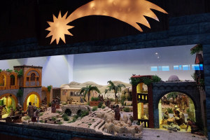 Eine Krippenausstellung im Museum mit Figuren, Tieren, Gebäuden und Grünpflanzen, beleuchtet von einer Deckenlampe an der Wand.