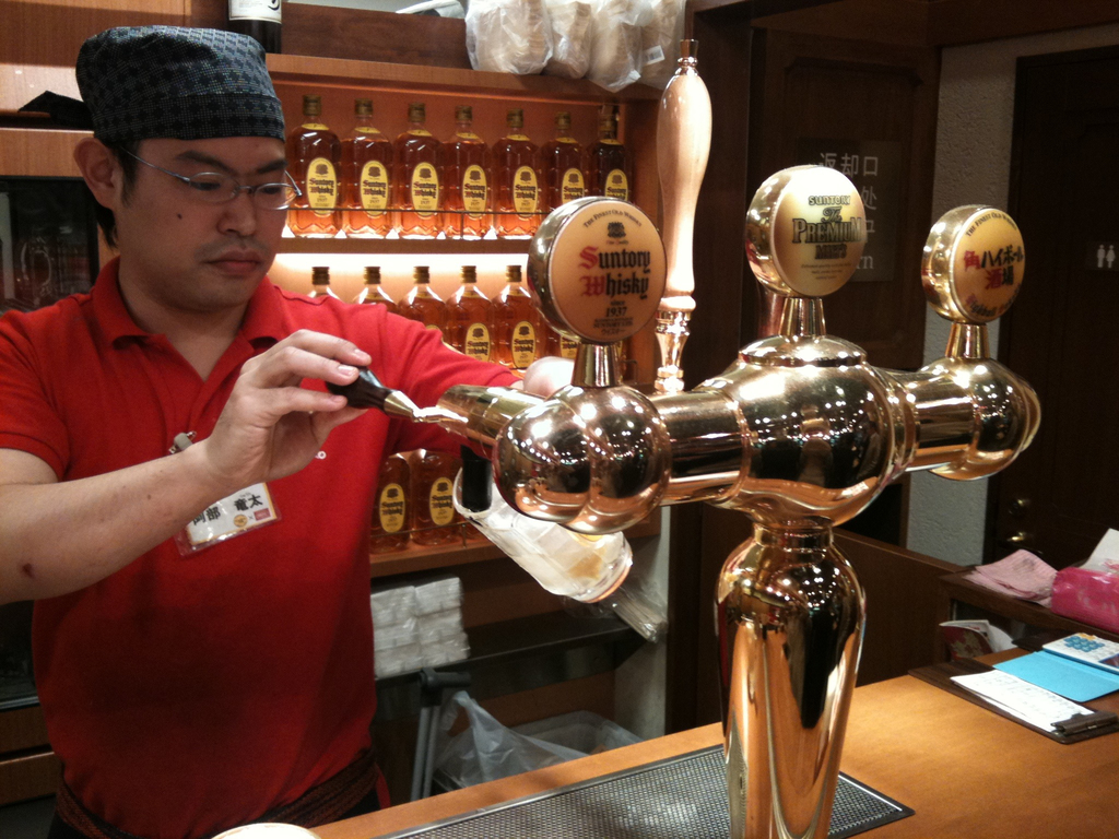 Eine Person mit M√ľtze und Brille f√ľllt ein Glas mit Bier von einem Automaten auf einem Tisch mit Papieren, mit Regalen voller Alkohol im Hintergrund.