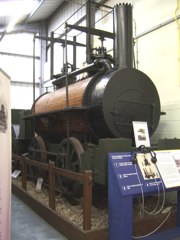 Alte Dampflokomotive in einem Museum ausgestellt mit Informationsschildern, einem Zaun, Steinen, einer Fahne, einer Wand mit Rohren und Deckenleuchten.