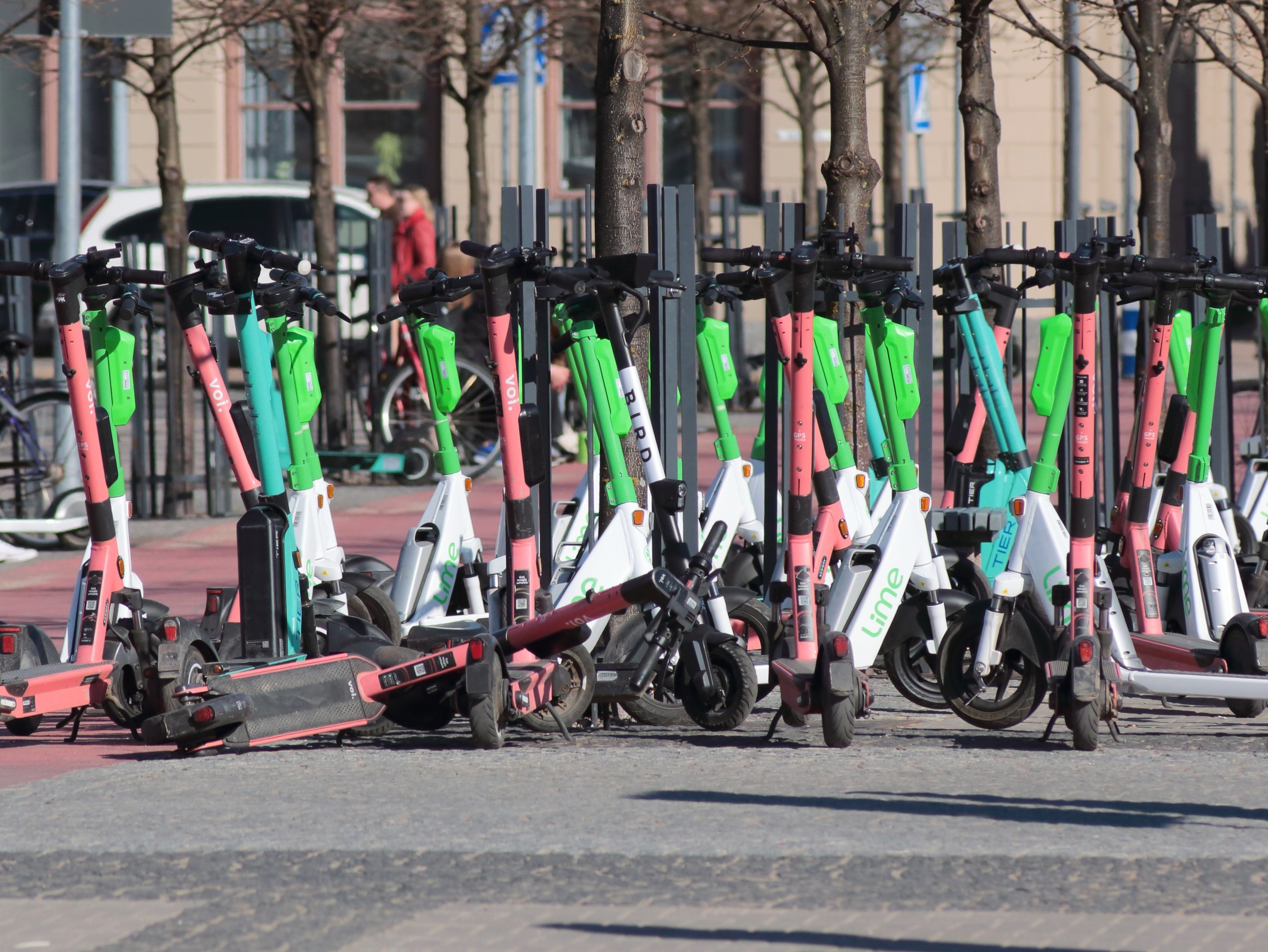 Eine Reihe von Elektro-Rollern auf einer Straße geparkt, mit einer Person im Vordergrund und unscharfen Hintergrundelementen wie Bäumen, Masten, Plakaten, Fahrzeugen und Gebäuden.