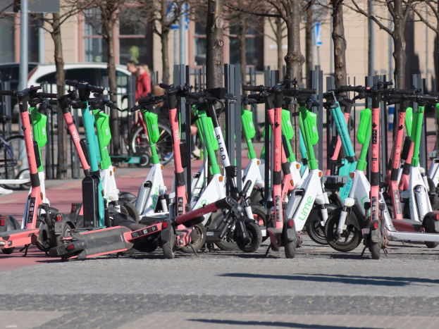 Eine Reihe von Elektro-Rollern auf einer Straße geparkt, mit einer Person im Vordergrund und unscharfen Hintergrundelementen wie Bäumen, Masten, Plakaten, Fahrzeugen und Gebäuden.