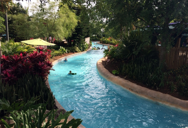 Ein Schwimmbad mit schwimmenden Menschen, umgeben von Pflanzen, Bäumen und einem Zaun, mit Menschen, Schirmen und Gebäuden im Hintergrund und Pfählen auf beiden Seiten.