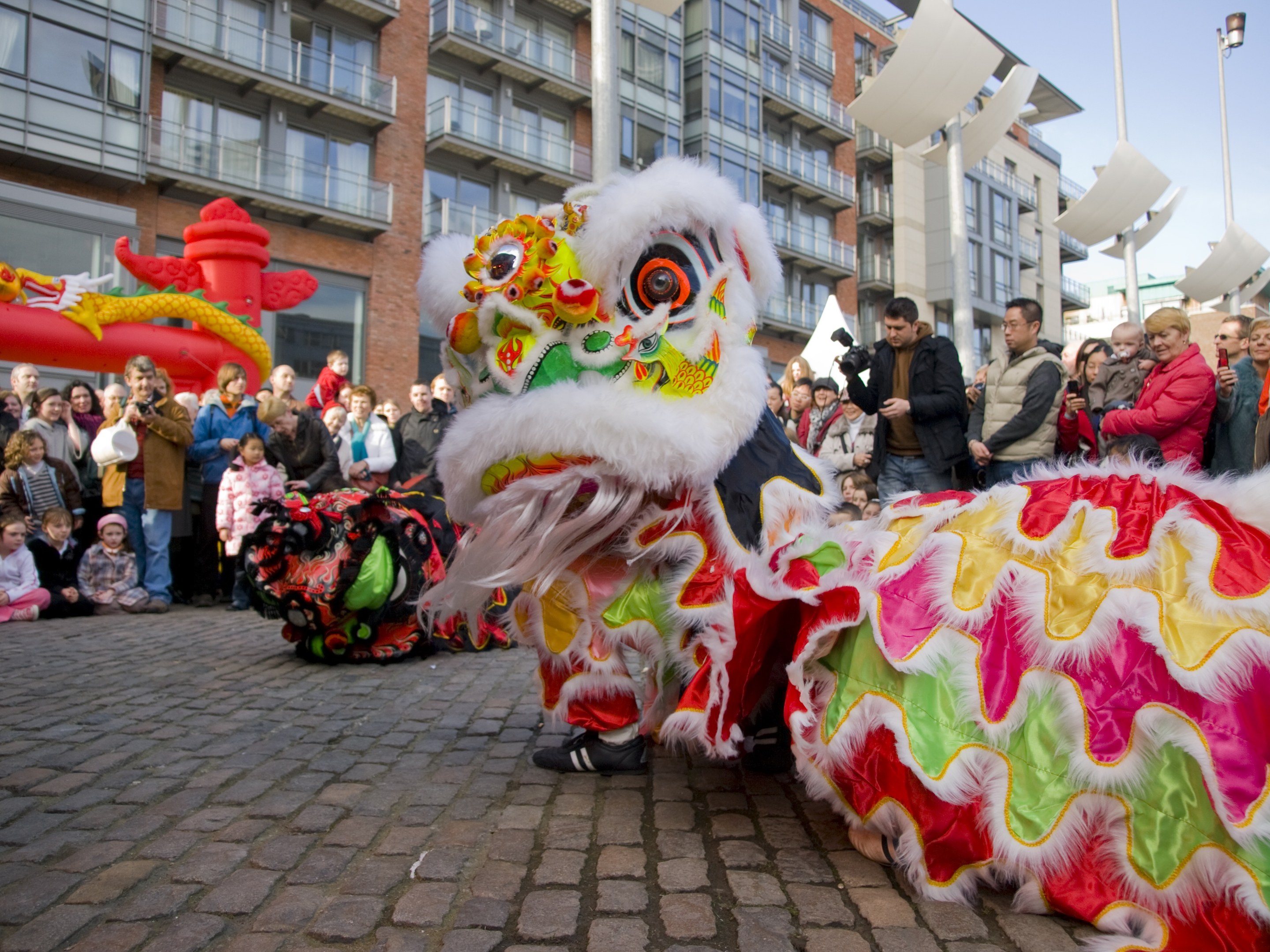 Ein lebendiges chinesisches Neujahrsfest in Amsterdam mit einer Löwen-Tanz-Show vor einem Publikum, das Kameras hält, vor Gebäuden und Laternen unter einem klaren blauen Himmel.