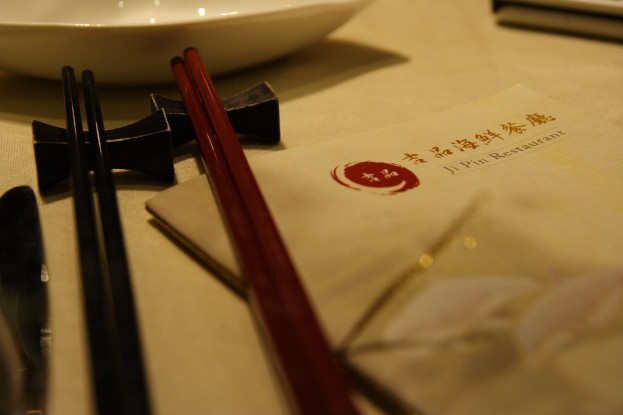 Ein Tisch mit einer Speisekarte, die 'Ji Pin Restaurant' auf Mandarin und Englisch lautet, sowie zwei Paar Stäbchen (eines rot, eines schwarz) und eine weiße Schüssel vor den Stäbchen.