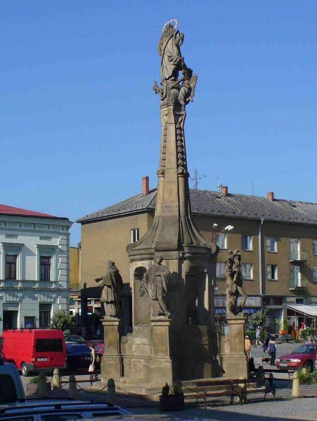 Ein Denkmal steht in einem Stadtplatz umgeben von Gebäuden, Straßenmöbeln, Fahrzeugen, Fußgängern, Bänken, Pflanzen und einem bewölkten Himmel.