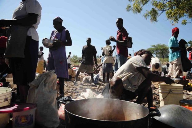 Eine Gruppe von Menschen, die in einer ländlichen Umgebung Essen zubereiten, mit einem großen Topf, Eimern und anderen Gegenständen auf dem Boden, unter einem klaren blauen Himmel.