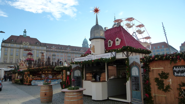 Ein belebter Weihnachtsmarkt in einem Stadtplatz mit geschmückten Bäumen, Gebäuden, Fahrzeugen, Passanten, Straßenlaternen und einem bewölkten Himmel.