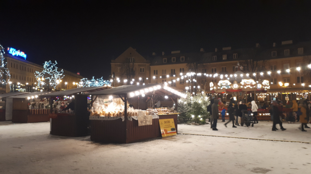 Lebendiger Weihnachtsmarkt bei Nacht auf einer verschneiten Straße mit Menschen, Buden mit Lichtern, Pflanzen, Bäumen, Gebäuden, Schildern und einem bewölkten Himmel.