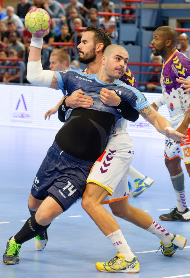 Männer spielen drinnen Handball, einer hält den Ball, mit Zuschauern auf Stühlen, Geländern und einer Wand in einer Stadionatmosphäre.