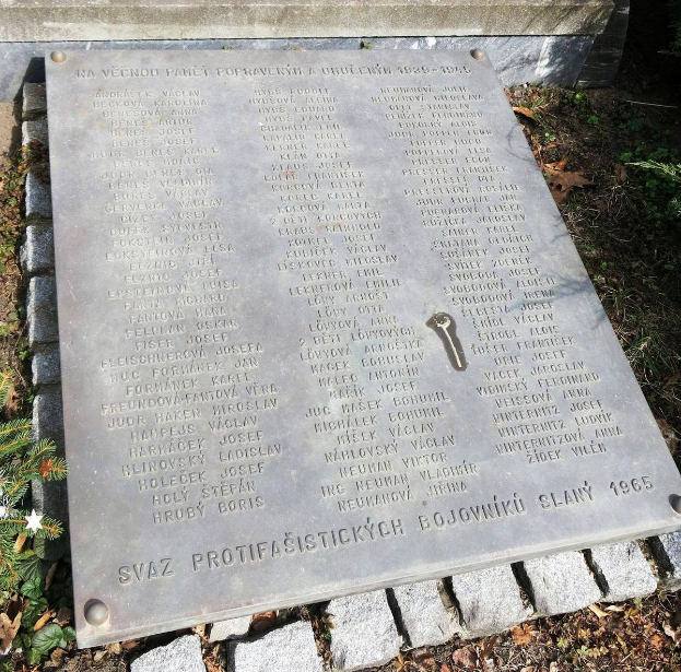 Ein Steinplaque an einer Wand in einem Park, umgeben von Pflanzen und trockenen Blättern, gedenkt den Opfern des Holocaust mit eingravierten Text.
