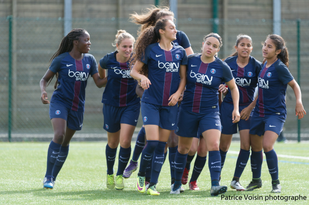 Gruppe junger Frauen beim Fußballspielen auf einem Rasenfeld mit Maschendrahtzaun und einer Wand im Hintergrund, Text in der Ecke lautet "Paris Saint-Germain Frauenfußball".