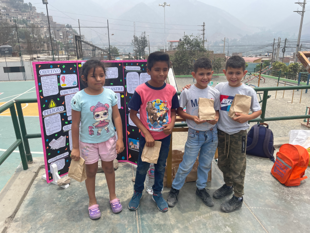 Eine Gruppe von Kindern steht auf einem Basketballfeld, jedes hält einen Papiertüte, mit Vorräten und einer Texttafel links und einem eingezäunten Bereich, Pfosten, Bäumen, Gebäuden und Hügeln im Hintergrund.