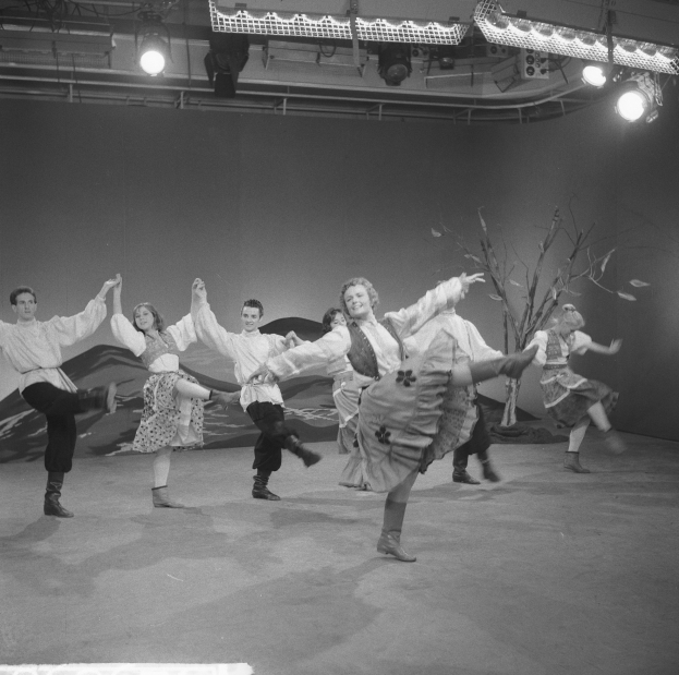 Eine Gruppe von Menschen, die auf einer Bühne tanzen, beleuchtet von Deckenlampen und umgeben von einer Wand, dargestellt in Schwarz-Weiß.