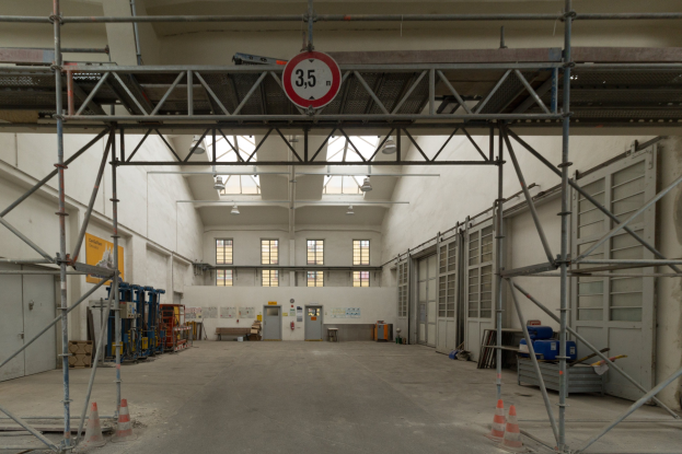 Lagerinneres mit Gerüsten, Verkehrskegeln, Metallstangen, einem Schild, Maschinen, einer Tür, Fenstern, Wänden, Deckenleuchten und einem Dach.