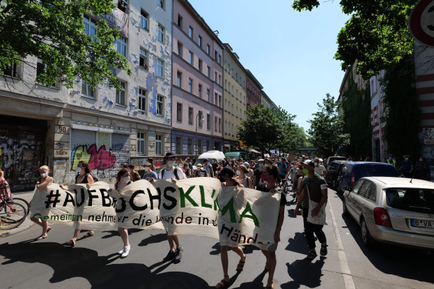 Eine Gruppe von Menschen marschiert mit einem Spruchband die Straße entlang, mit parkenden Autos auf der rechten Seite, einem Fahrrad auf der linken Seite und Gebäuden, Bäumen und einem Schild im Hintergrund unter einem sichtbaren Himmel.