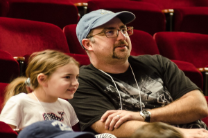 Ein Mann mit Brille, Uhr und Mütze sitzt mit einem strahlenden kleinen Mädchen in einem Kino, umgeben von anderen Menschen und Stühlen.