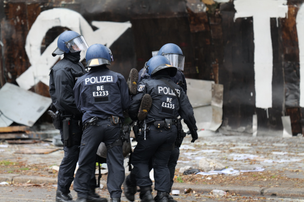 Polizisten in schwarzen Uniformen und blauen Helmen stehen vor einem mit Graffiti bedeckten Gebäude während einer Unruhen, mit verstreuten Papieren und Gegenständen auf dem Boden.