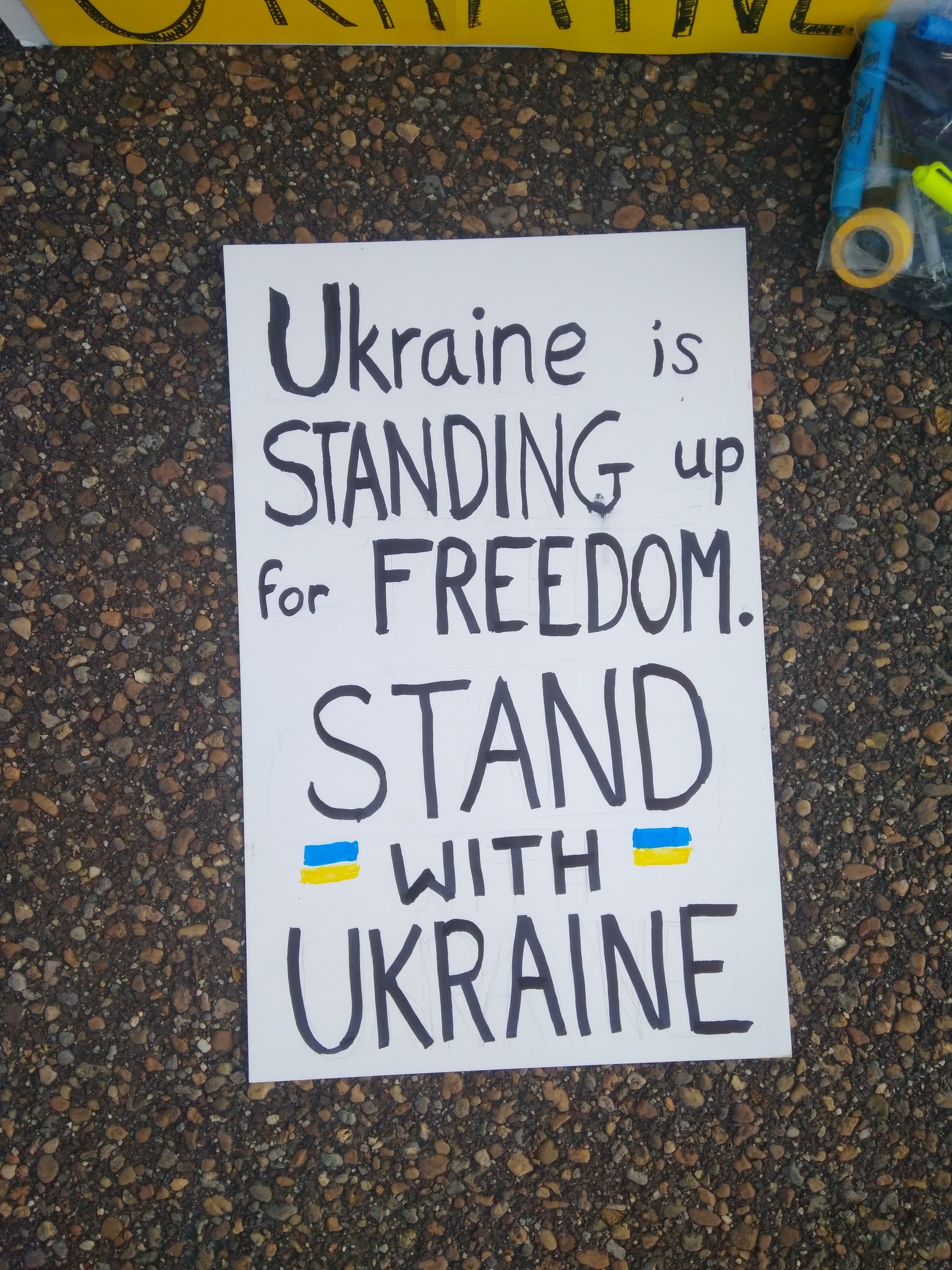 Weißer Zettel mit dem Text "Ukraine steht für Freiheit ein, steh mit Ukraine" auf dem Boden vor einem gelben Banner, mit einer Plastikabdeckung mit verschiedenen Gegenständen rechts.