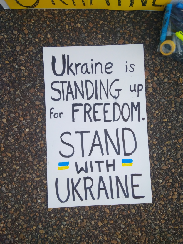 Weißer Zettel mit dem Text "Ukraine steht für Freiheit ein, steh mit Ukraine" auf dem Boden vor einem gelben Banner, mit einer Plastikabdeckung mit verschiedenen Gegenständen rechts.