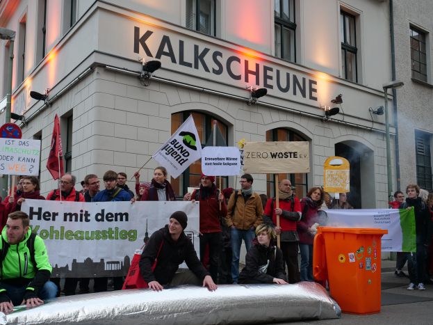Eine Gruppe von Menschen mit Transparenten und Schildern steht vor einem Gebäude, mit zwei Personen im Vordergrund und einem Müllcontainer rechts daneben, während einer Demonstration in Deutschland.
