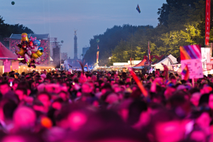 Eine große Menschenmenge bei einem nächtlichen Musikfestival, beleuchtet von bunten Lichtern, mit Zelten, Fahnen, Masten und anderen Gegenständen im Hintergrund und einem Turm, Bäumen und einem klaren Nachthimmel