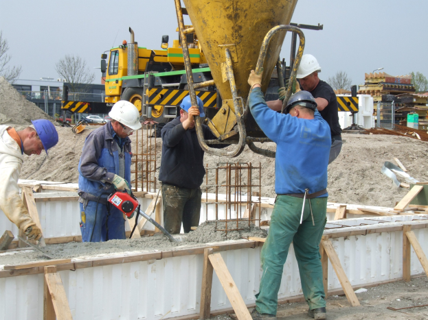 Männer mit Helmen bedienen einen Betonmischer auf einer Baustelle mit Fahrzeugen, Bäumen, Gebäuden und einem klaren blauen Himmel im Hintergrund.