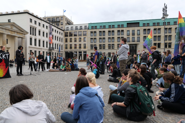 Eine Gruppe von Menschen, die auf dem Boden vor einer Menge mit Fahnen und Spruchbändern sitzt, während einer Anti-Schwulen-Demo in Berlin. Im Hintergrund sind eine Statue, ein Mikrofonständer und Gebäude zu sehen.