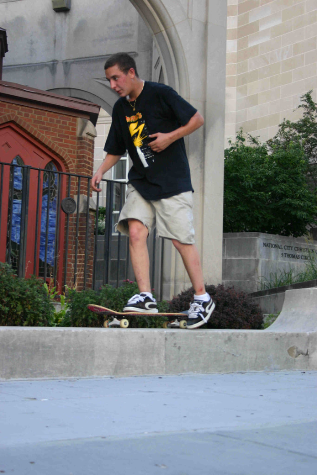 Ein Mann fährt Skateboard auf einer vertikalen Wand mit einer Plattform unten, ein Haus, Grill, Pflanzen, Bäume, eine Backsteinwand und eine Säule im Hintergrund.