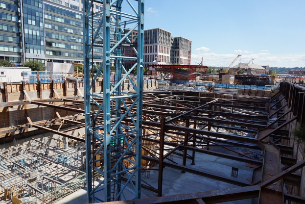 Das Bild zeigt eine Baustelle mit Gebäuden und Büros links, Eisenstangen und Maschinen rechts und verschiedene Ausrüstungen und Kräne in der Mitte, unter einem Himmel in der oberen rechten Ecke.