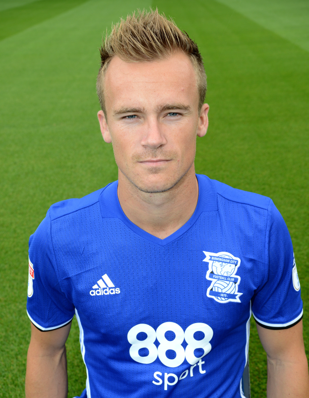 Ein Mann in einem blauen T-Shirt steht selbstbewusst auf einem Fußballfeld und sieht aus wie ein Flügelspieler mit entschlossener Miene.