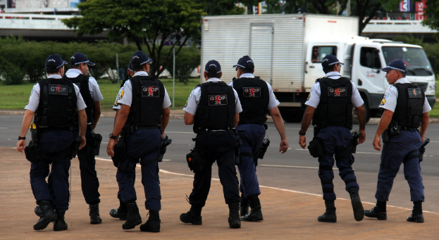 Eine Gruppe von Beamten in Uniform, einschließlich Mützen, Hemden, kugelsicheren Westen, Uhren, Hosen und Schuhen, die auf einer Straße mit einem geparkten Lastwagen auf der rechten Seite neben einem Zaun, Gebäuden, Bäumen, Pflanzen, Pfählen und Bannern im Hintergrund und einer Brücke oben rechts gehen.