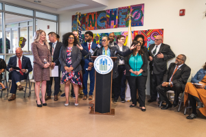 Gruppe von Menschen in einem Raum mit einem Podium, einem Mikrofon und einer Tafel mit der Aufschrift "Sancho-Stadtrat tritt dem Stadtrat bei", umgeben von Stühlen, Flaggen, einem Feuerlöscher und Deckenleuchten.
