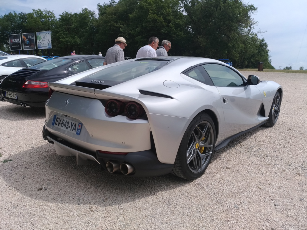 Roter Ferrari 812 Superfast auf dem Goodwood Festival of Speed mit Zuschauern in Mützen, Bäumen, Infotafeln und bewölktem Himmel im Hintergrund.
