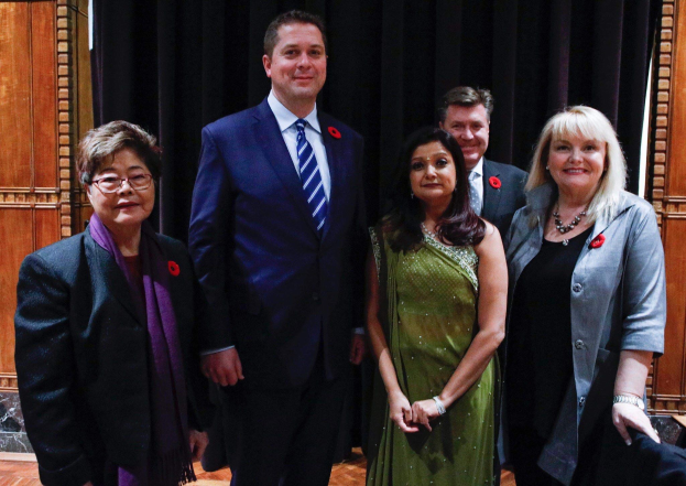 Gruppe von Menschen, die vor einem schwarzen Vorhang mit einer hölzernen Wand im Hintergrund stehen und wie Mitglieder der Liberalen Partei auf einer Versammlung aussehen.