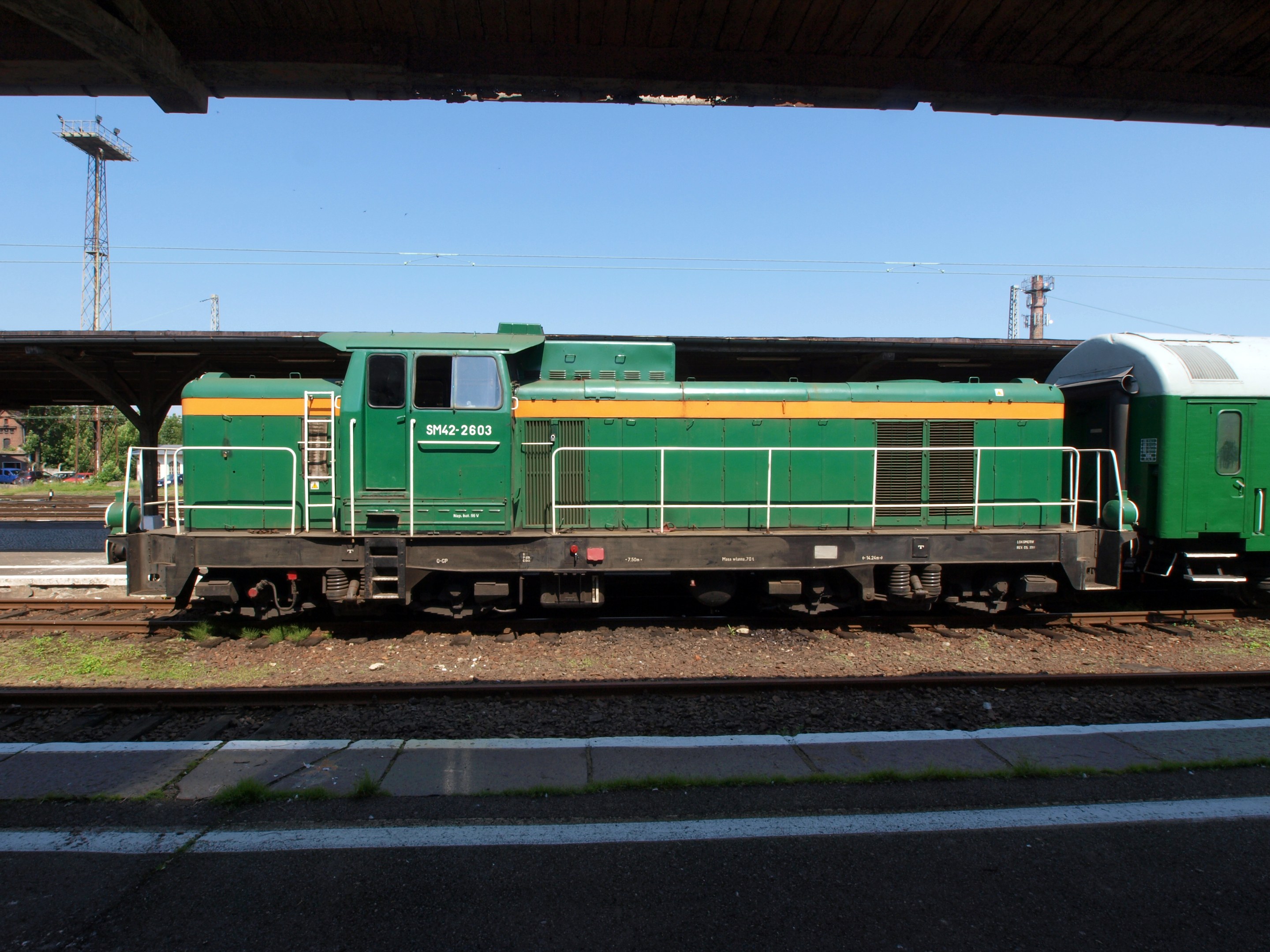 Grüne Diesel-Elektrolokomotive auf Eisenbahnschienen unter einer Brücke, umgeben von Steinen, Gras, einem Weg, Bäumen, Strommasten mit Drähten, einem Turm, Fahrzeugen und einem bewölkten Himmel.