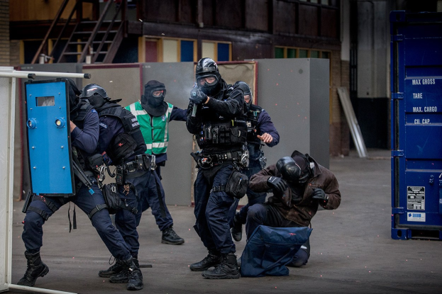 Polizeibeamte in Einsatzausrüstung stehen vor einem Gebäude in einer Reihe, einige halten Waffen, mit einer Person auf dem Boden im Vordergrund und einem blauen Objekt mit Text auf der rechten Seite.