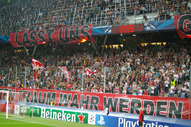 Ein Fußballspiel in einem Stadion mit einer großen Menge, saftigem Grün, einem Torpfosten links, Bodenbanner und Fahnen haltenden Menschen.