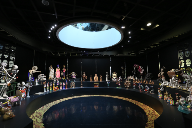 Ein großer Raum voller verschiedener Spielzeuge, darunter Skelette, die auf dem Boden angeordnet sind, mit Totenschädeln an den Wänden und Lampen an der Decke, die eine festliche Atmosphäre für den Tag der Toten schaffen.