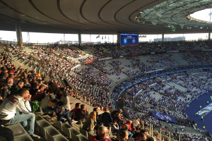 Eine große Menschenmenge sitzt im Allianz Arena Stadion in München, Deutschland, bei einem Fußballspiel, mit einer Bühne auf der rechten Seite, Fahnen, Stangen und einem Bildschirm im Hintergrund, unter einem sichtbaren Himmel.