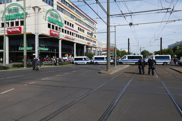 Gruppe von Polizeibeamten steht auf der Straße neben einer Straßenbahn, mit Fahrzeugen, Strommasten, Gebäuden, Bäumen und einem bewölkten Himmel im Hintergrund.
